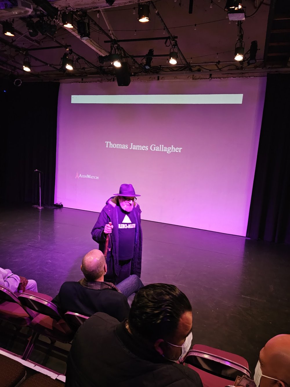 A reader at the podium at Highways, with a World AIDS Day title card on screen behind him.