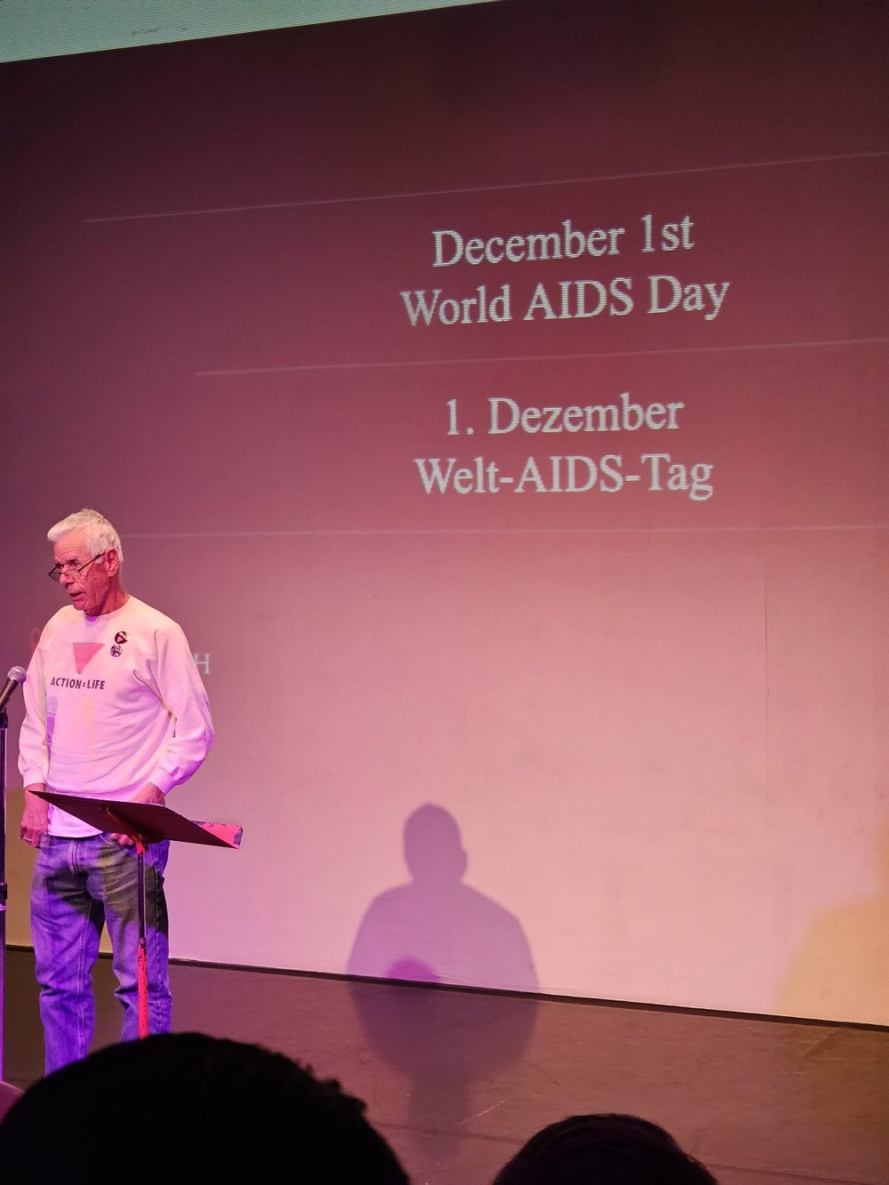 A reader at the podium at Highways under purple lighting, with an AIDSWatch name on screen behind him.