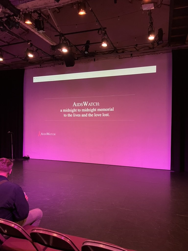 AIDSWatch projected on the full screen at Highways, reading 'a midnight to midnight memorial to the lives and the love lost,' with an audience member seated below.